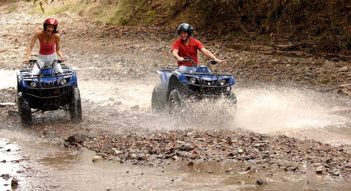 Zwei Leute auf Quads im Wasser, einer mit rotem Helm und roter Shirt, die andere mit rotem Helm und weißem Shirt.