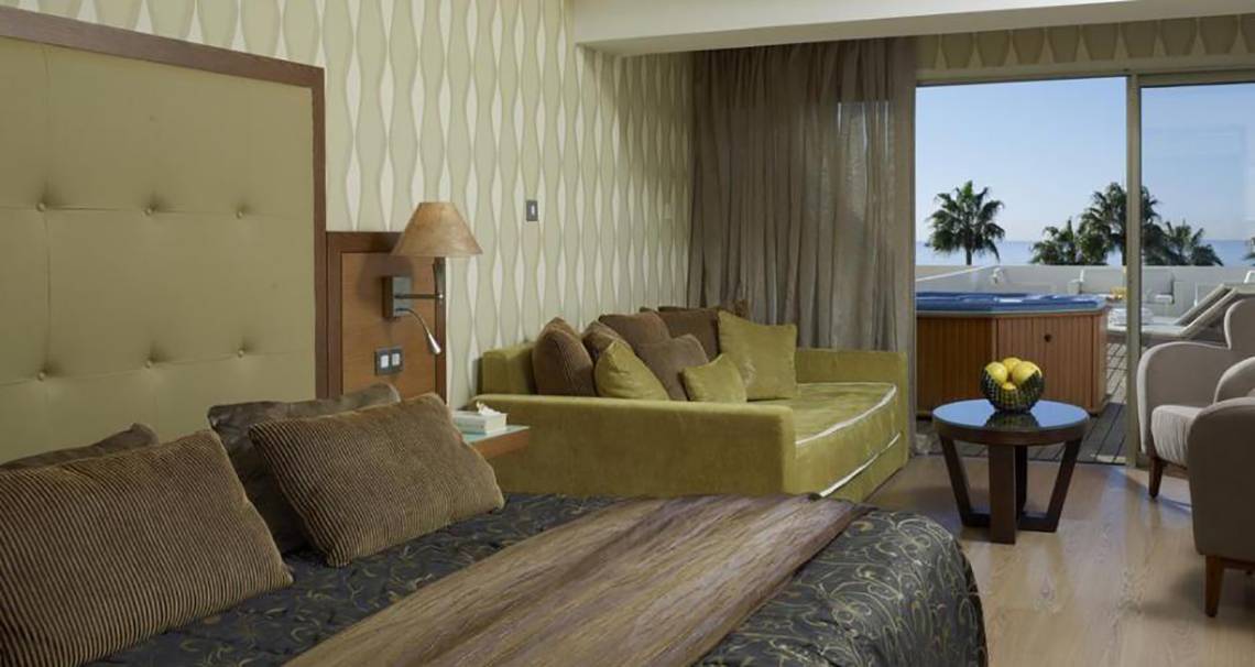 Hotelzimmer im Atlantica Miramare Beach Hotel mit großem Bett, Sofa, Tisch und Whirlpool auf dem Balkon.