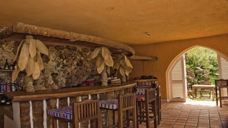 Innenansicht der Bar im Bahari Beach Hotel mit einer Steinmauer, Holzbar, Stühlen und einer Aussicht auf den Garten.