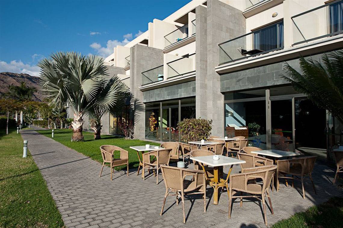 Terrasse des LIVVO Costa Taurito Hotels mit Tischen und Stühlen, umgeben von Palmen und Gras.