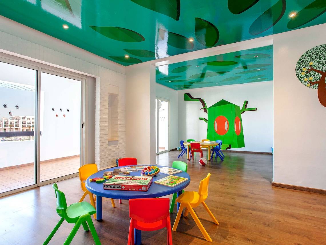 Kinderspielzimmer im Bianca Beach Resort mit bunten Tischen und Stühlen, blauem Tisch, grüner Wandmalerei und bemalter Decke.