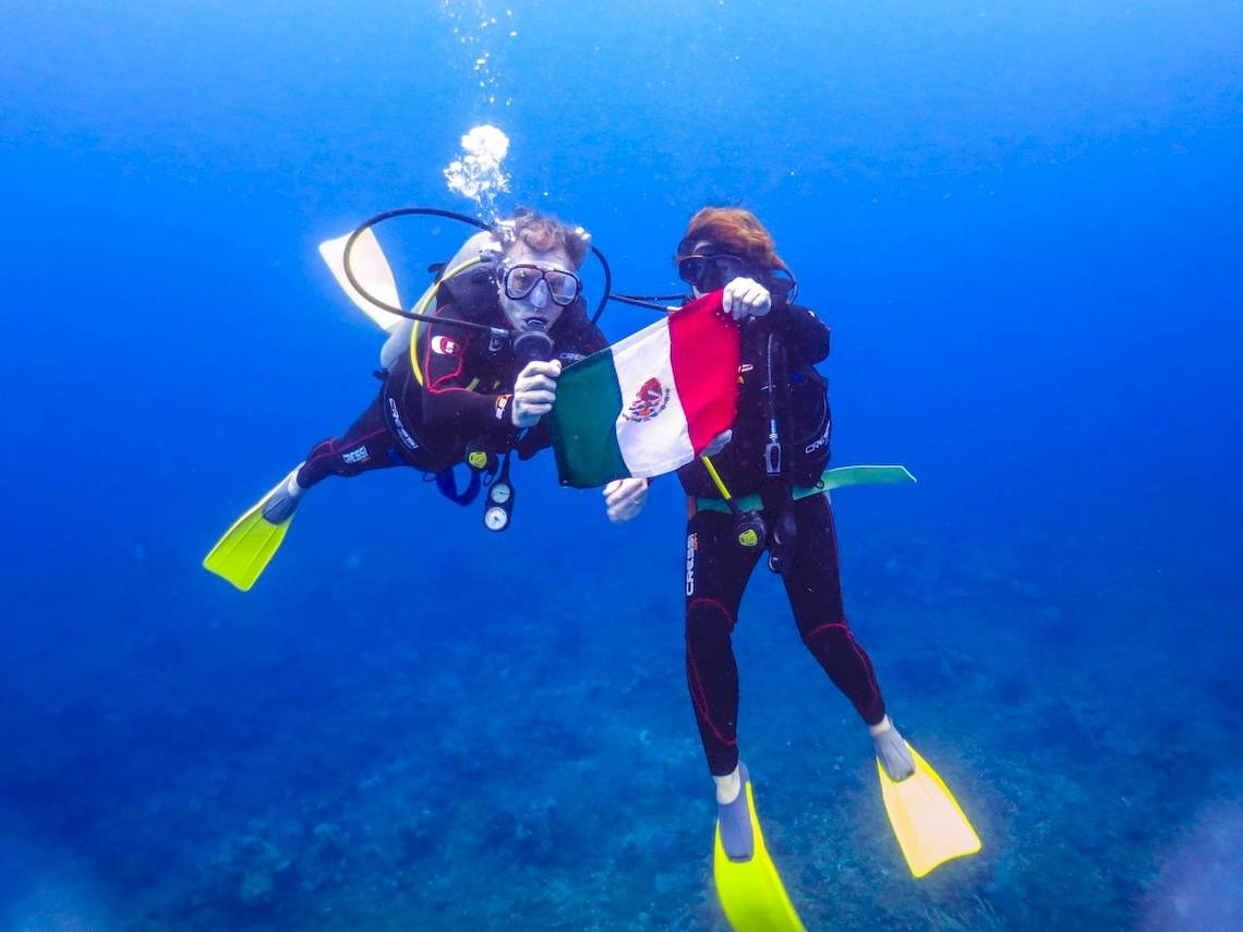 Zwei Taucher im Wasser halten eine mexikanische Flagge vor sich