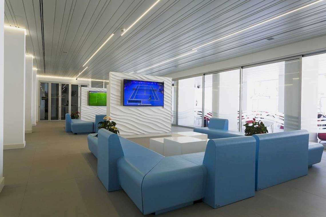 Lobby-Lounge-Bereich des Caserio Hotels mit blauen Sofas, Tischen und Fernsehern.