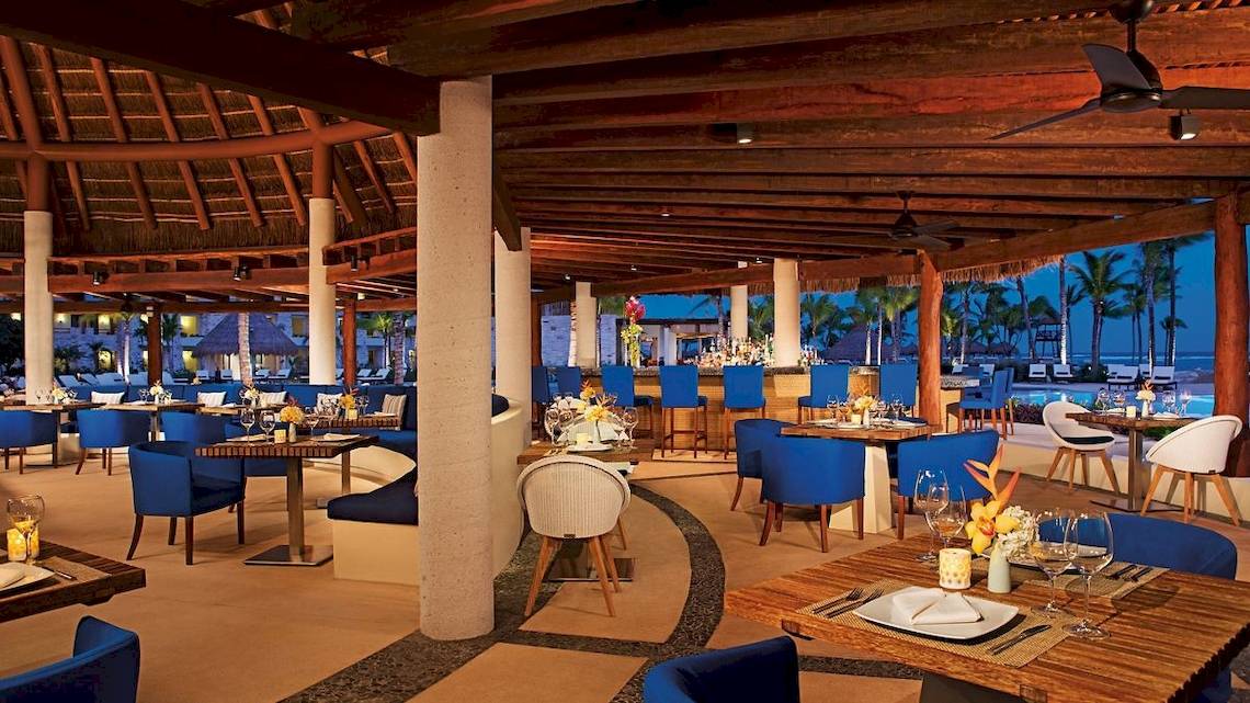Innenbereich des Restaurants im Secrets Akumal Riviera Maya mit blauen Stühlen und Tischen, Holztischen, Deckenventilatoren und Blick auf den Ozean.