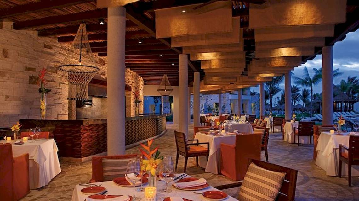 Innenbereich des Secrets Akumal Riviera Maya Hotels mit rustikalem Design, Säulen, Beleuchtung und gedeckten Tischen.