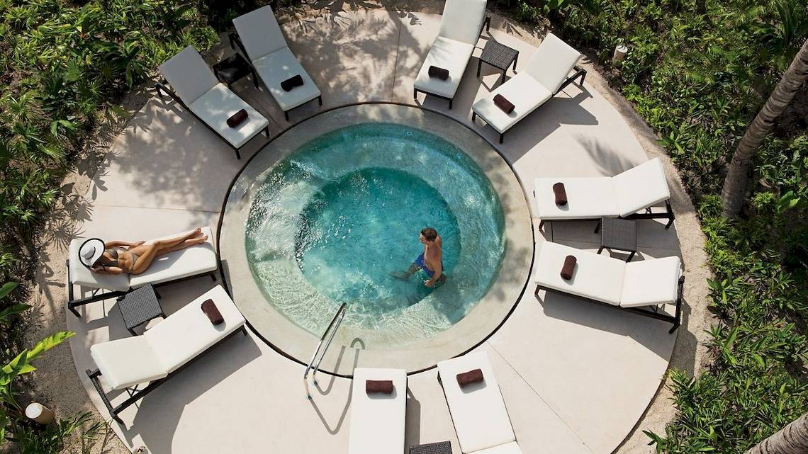 Oberflächenansicht eines Infinity-Pools im Secrets Akumal Riviera Maya Hotel mit mehreren weißen Liegestühlen und grüner Umgebung.