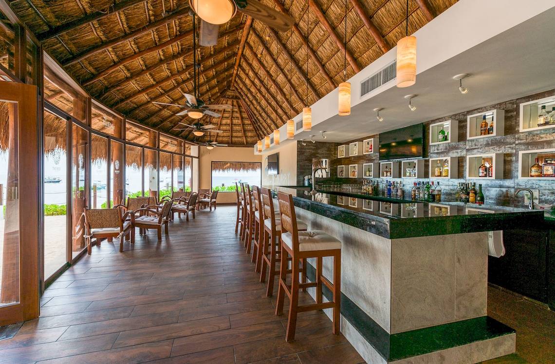 Innenansicht der Bar im Reef Coco Beach Hotel mit Barhockern, Theke und Fenstern.