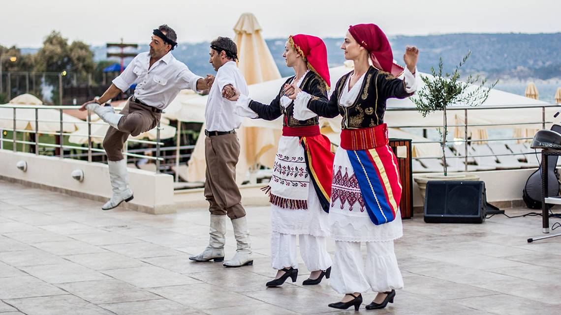 Vier Personen in traditioneller griechischer Kleidung tanzen auf der Terrasse des Kiani Beach Resorts.