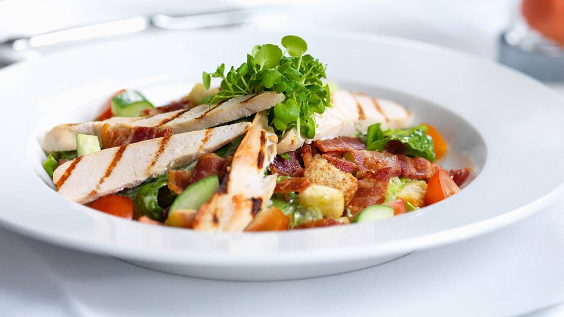 Ein Gericht im weißen Teller mit Salat, gegrilltem Fleisch, Tomaten und Gurken.