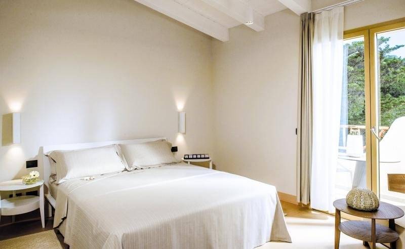 Schlafzimmer im Hotel Cala Cuncheddi mit einem Bett, Kissen, Tisch und Aussicht auf Bäume.