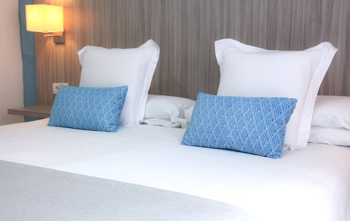 Eingemachtes Bett im O7 Aloe Corralejo Hotel mit weißen Laken, Kissen und blauen Musterkissen.