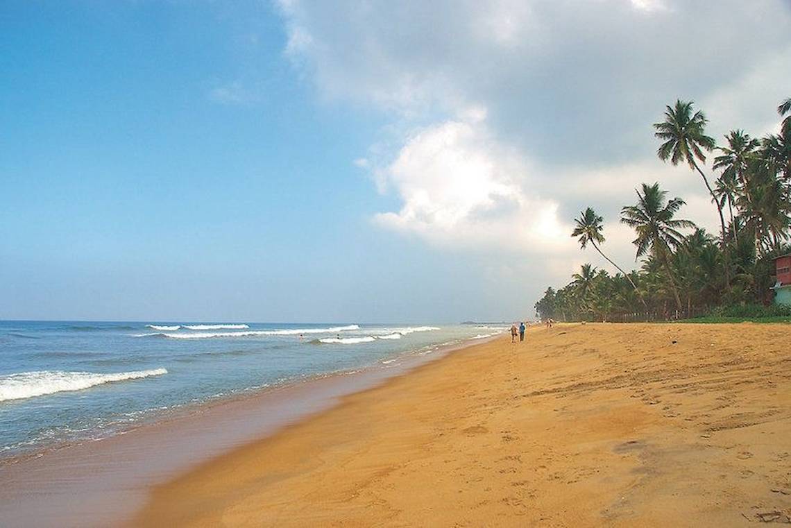 Strand mit Palmen und zwei Personen, die am Meer entlanggehen