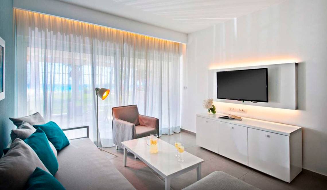 Wohnzimmer im Nissi Beach Resort mit Fernseher, Couch, Tisch, Stuhl und Lampe.