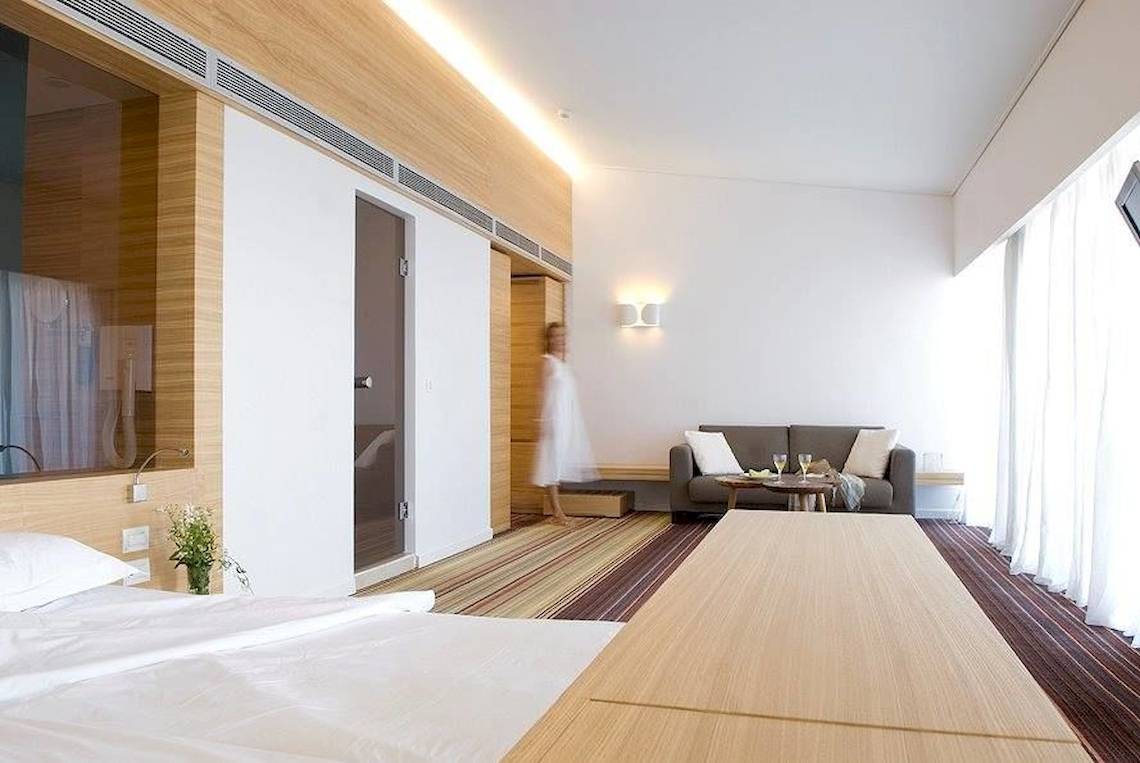 Innenansicht eines Hotelzimmers im Doryssa Seaside Resort, mit Bett, Wohnzimmerbereich und Frau im Hintergrund.