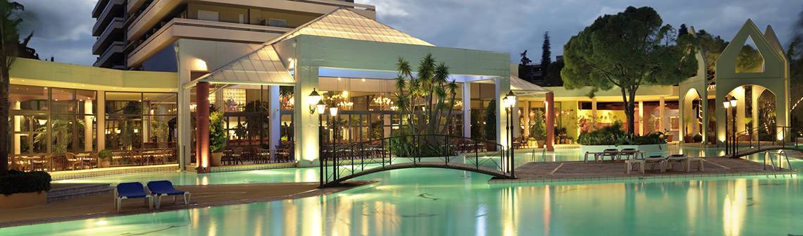Der Außenpool des Dionysos Hotels bei Nacht mit einer Brücke und Lounge-Stuhl am Pool.
