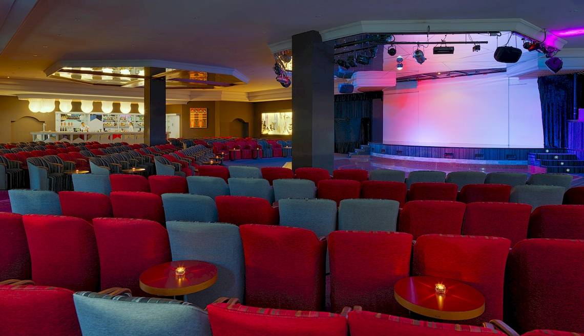 Innenansicht des H10 Rubicon Palace Hotels mit einem großen Saal, roten und grauen Stühlen, und einer Bühne.