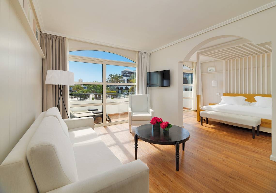 Innenansicht eines Zimmers im H10 Rubicon Palace Hotel mit Sofa, Tisch, Sessel und Fernseher.