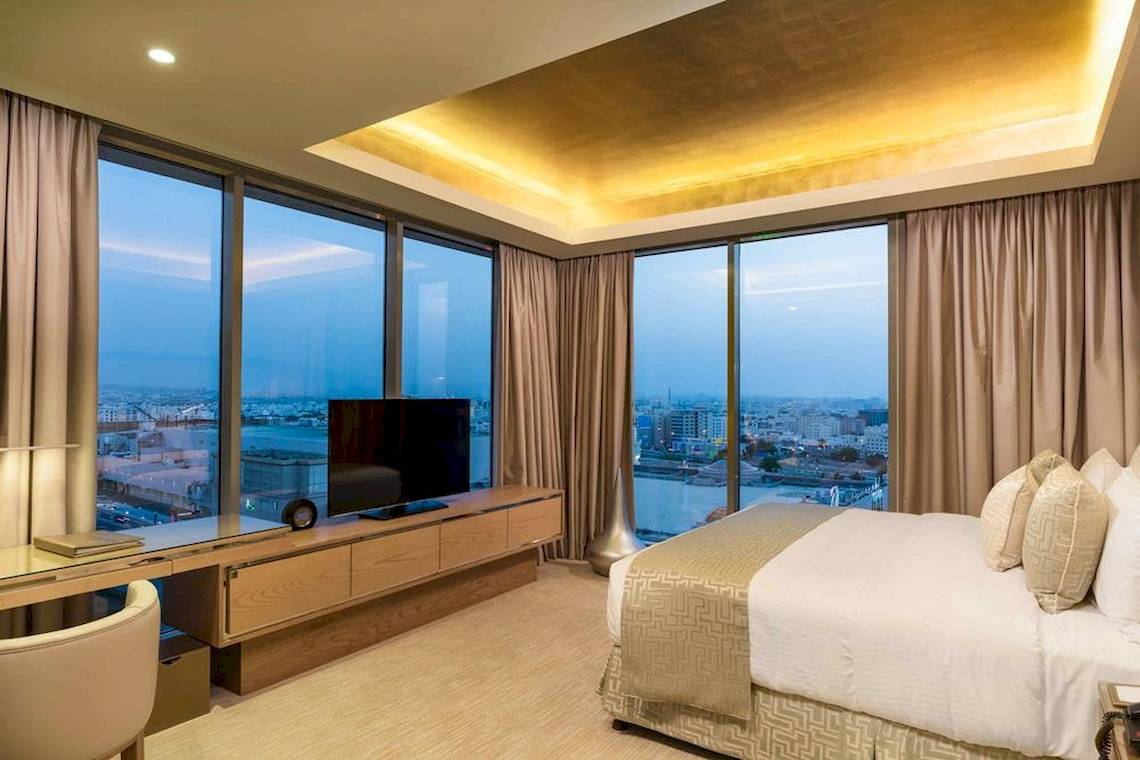 Innenansicht eines Hotelzimmers im Grand Millennium Muscat mit einem großen Bett und Stadtblick.