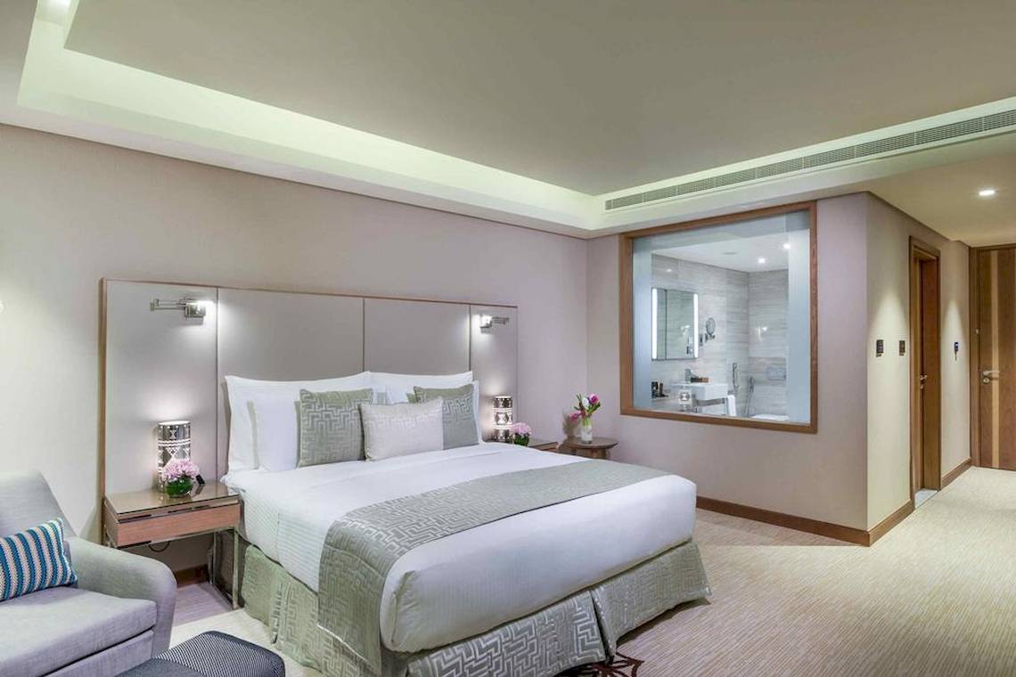 Elegantes Zimmer im Grand Millennium Muscat Hotel mit Kingsize-Bett, Nachttischen und Ausblick auf das Badezimmer.