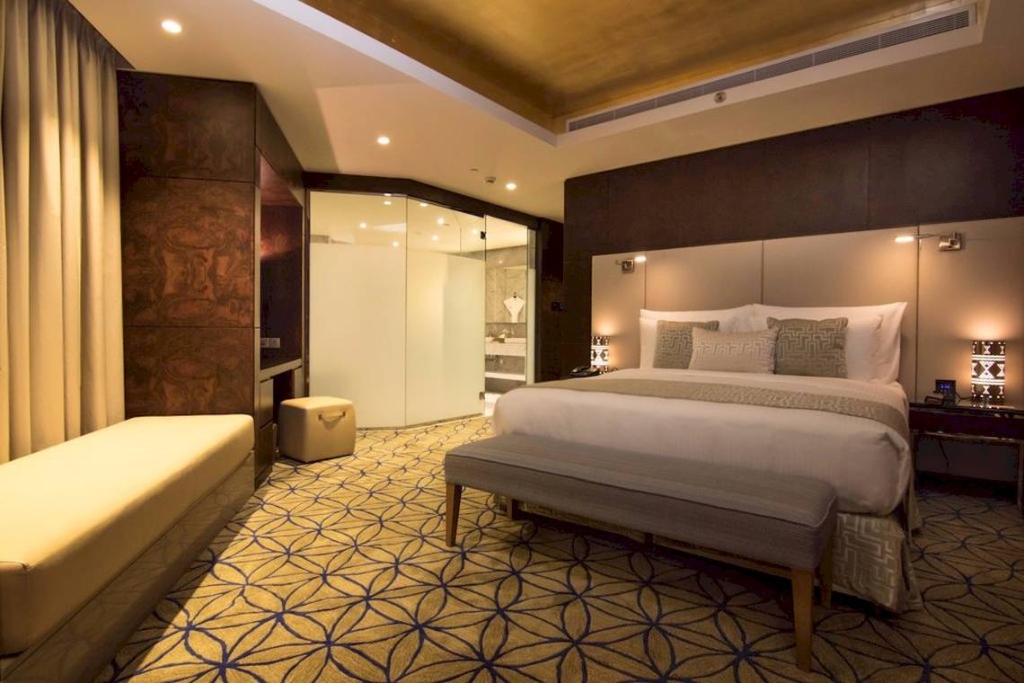 Innenansicht eines eleganten Schlafzimmers im Grand Millennium Muscat Hotel, mit großem Bett, gepolstertem Nachttisch und luxuriösem Badezimmer.