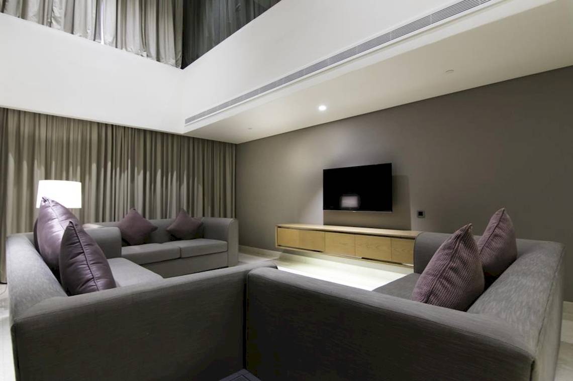 Wohnzimmer im Grand Millennium Muscat mit grauen Sofas, Kissen und Fernseher.