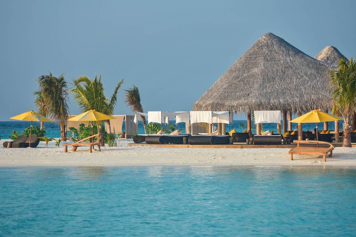 Strandszene des Drift Thelu Veliga Retreat mit Sonnenliegen, gelben Sonnenschirmen und einer großen runden Hütte
