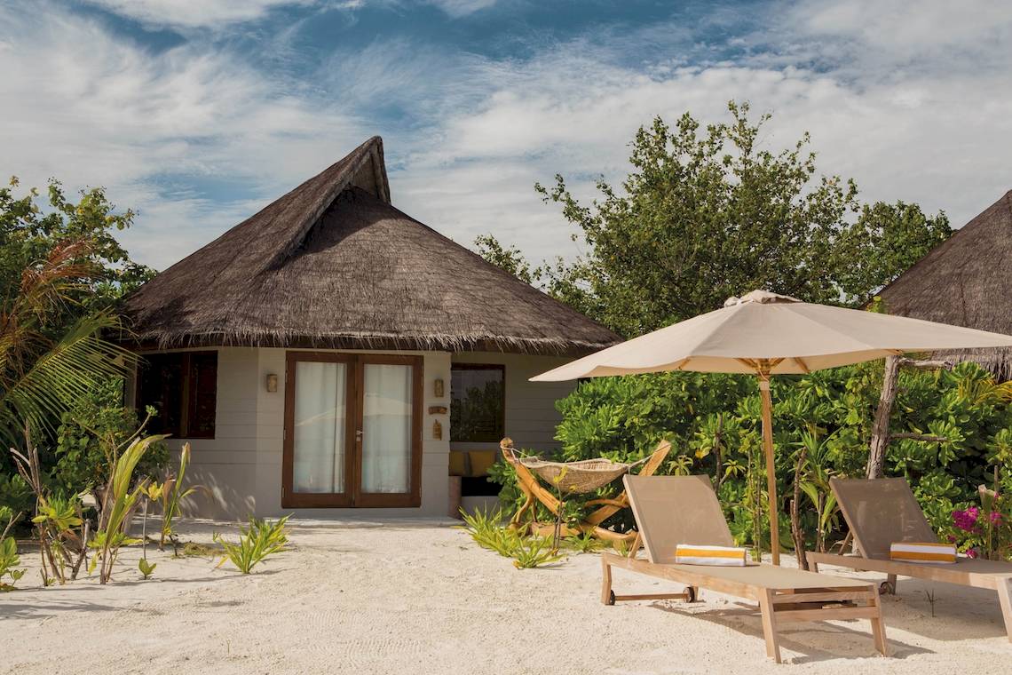 Außenansicht des Drift Thelu Veliga Retreat, eines kleinen, runden, strohgedeckten Häuschens am Strand mit einem Sonnenschirm und Liegestühlen davor.