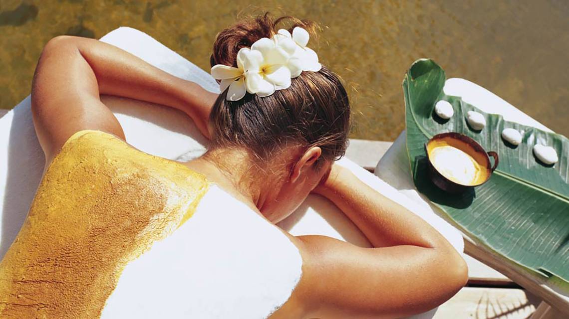 Rückenansicht einer Frau im Porto Elounda Golf & Spa Resort, die eine goldene Körperbehandlung genießt, mit einer weißen Blume im Haar und einem grünen Blatt neben einer Tasse.