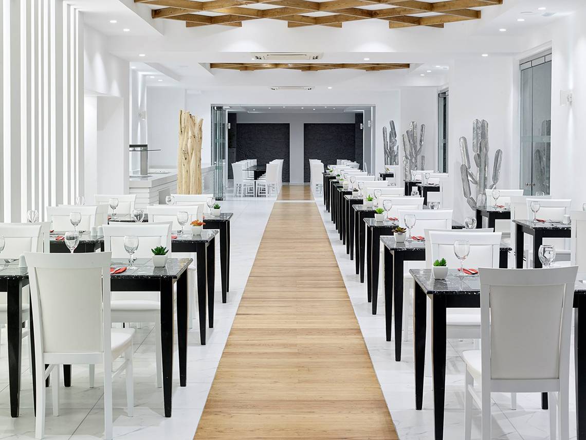 Innenansicht des Restaurants im Dimitra Beach Hotel & Suites mit weißen Tischen und Stühlen, einem hölzernen Gang und Kaktus-Dekorationen.