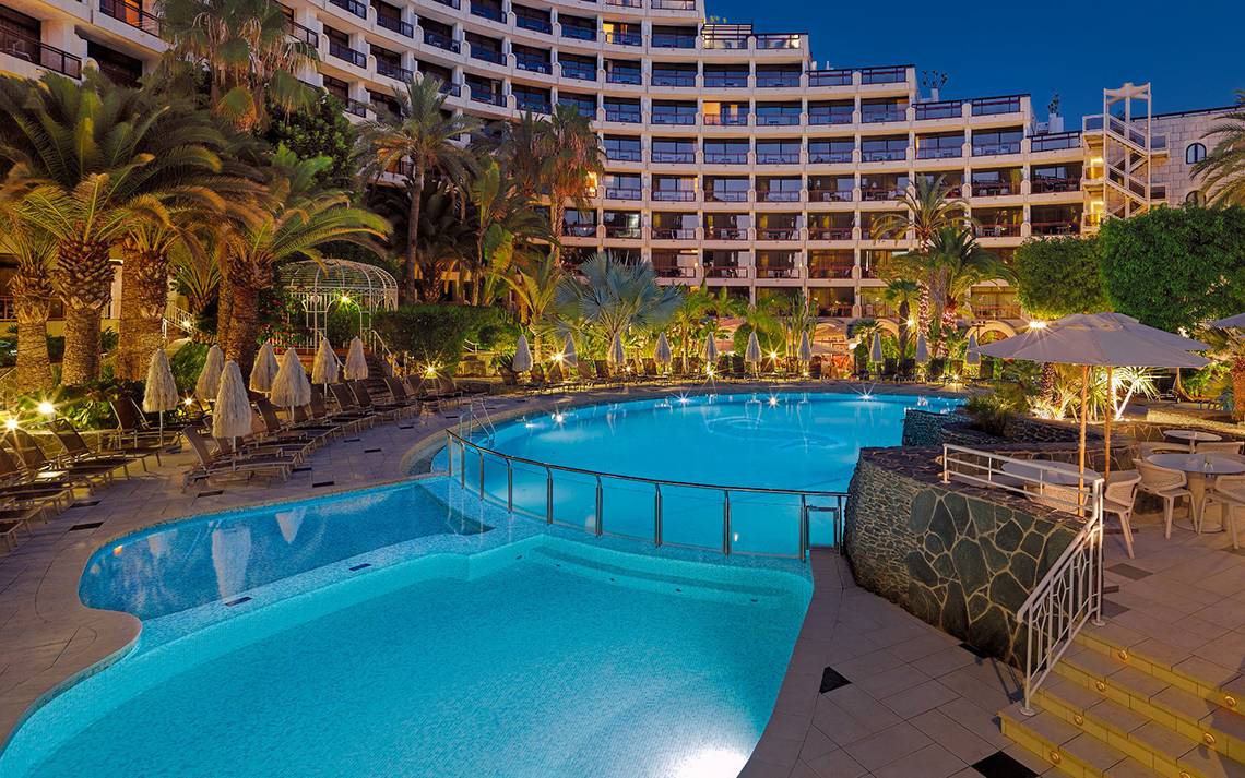 Abendansicht des Swimmingpools im Seaside Sandy Beach Hotel mit illuminierten Gebäuden und Sonnenliegen.