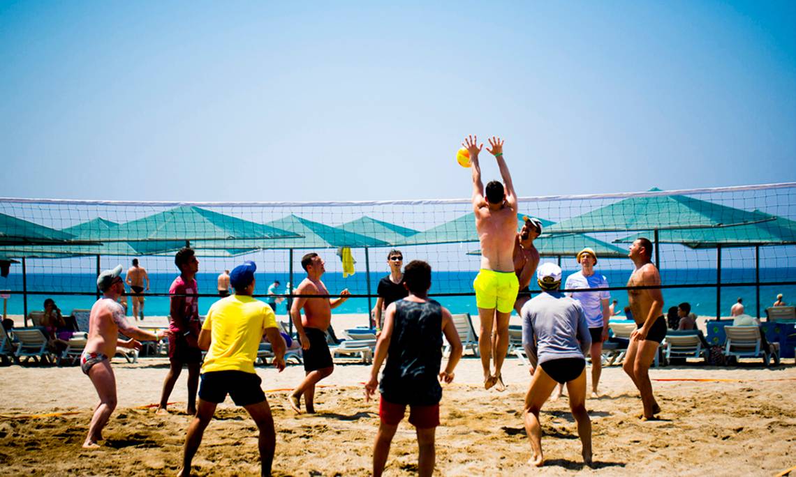 Mehrere Männer spielen Volleyball auf einem Strand mit einem Netz und Sonnenschirmen im Hintergrund.