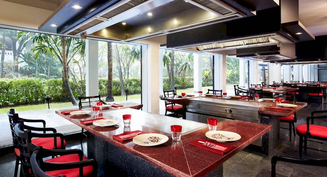 Innenbereich des Restaurants im Anantara Riverside Bangkok Resort mit roten Stühlen und Tischen, großen Fenstern und Blick auf Gärten