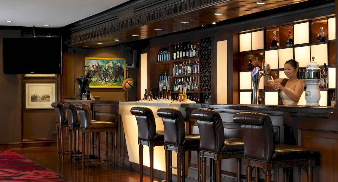 Hotelbar im Anantara Riverside Bangkok Resort mit Barkeeperin und voller Bar.