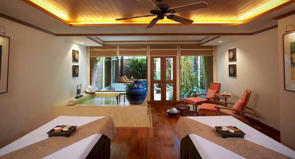 Massageraum im Anantara Riverside Bangkok Resort mit zwei Massagetischen, Blick auf einen Außenpool und Garten.