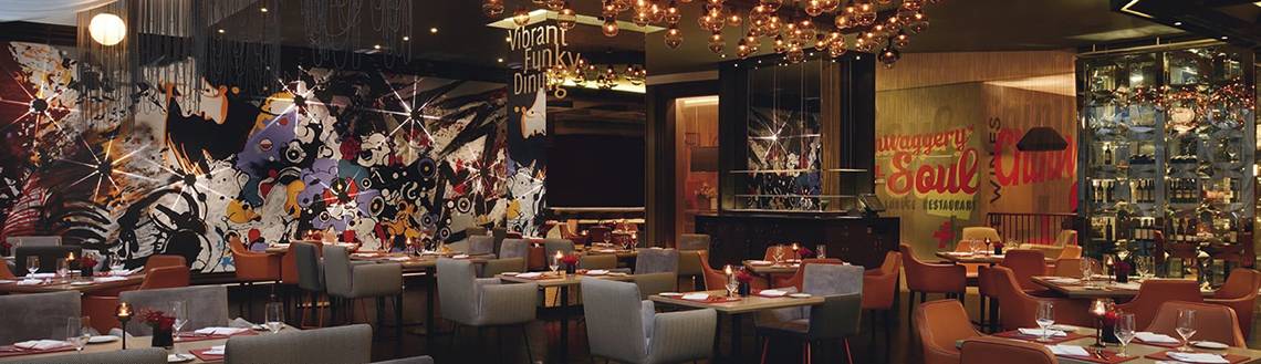 Innenraum des Mövenpick Hotel Jumeirah Beach Restaurant mit bunten Wandmalereien, Tischdekoration und hängenden Kronleuchtern.