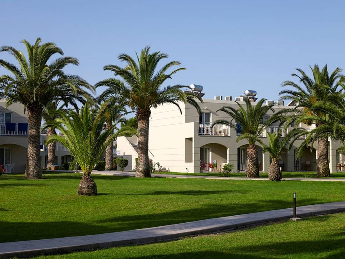Außenbereich des Atlantica Holiday Village Kos mit Gebäuden, Palmen und Rasen.