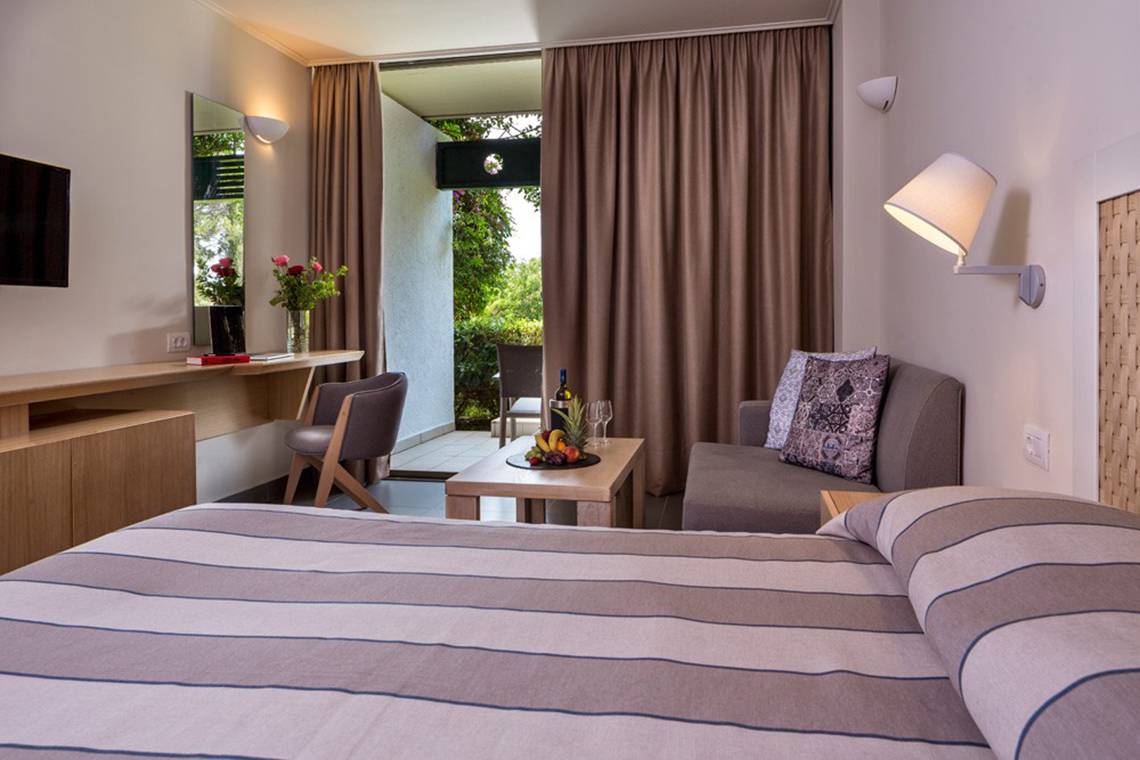 Innenansicht eines Hotelzimmers im Kontokali Bay Resort & Spa mit einem Bett, einem Sofa und einem Tisch mit Früchten.