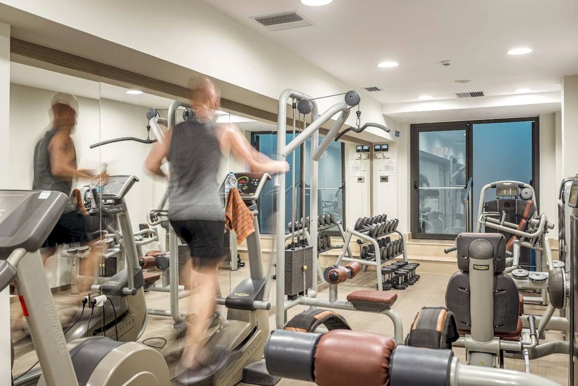 Fitnessstudio im Mitsis Rodos Village Beach Hotel mit verschiedenen Geräten und zwei Personen, die Cardiogeräte benutzen.