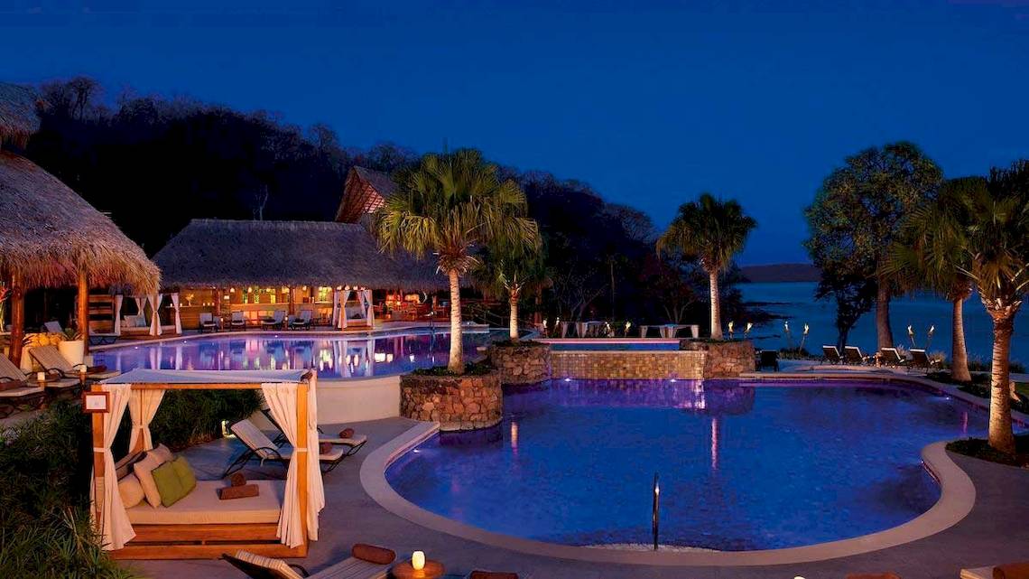 Nachtansicht des Secrets Papagayo Costa Rica Hotels mit beleuchtetem Infinity-Pool und Palmen.