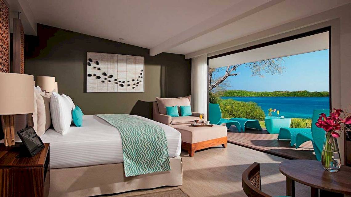 Suite-Zimmer mit Meerblick im Secrets Papagayo Costa Rica mit einem großen Bett, Kissen, Lampen und einem großen Fenster mit Blick auf das Meer.