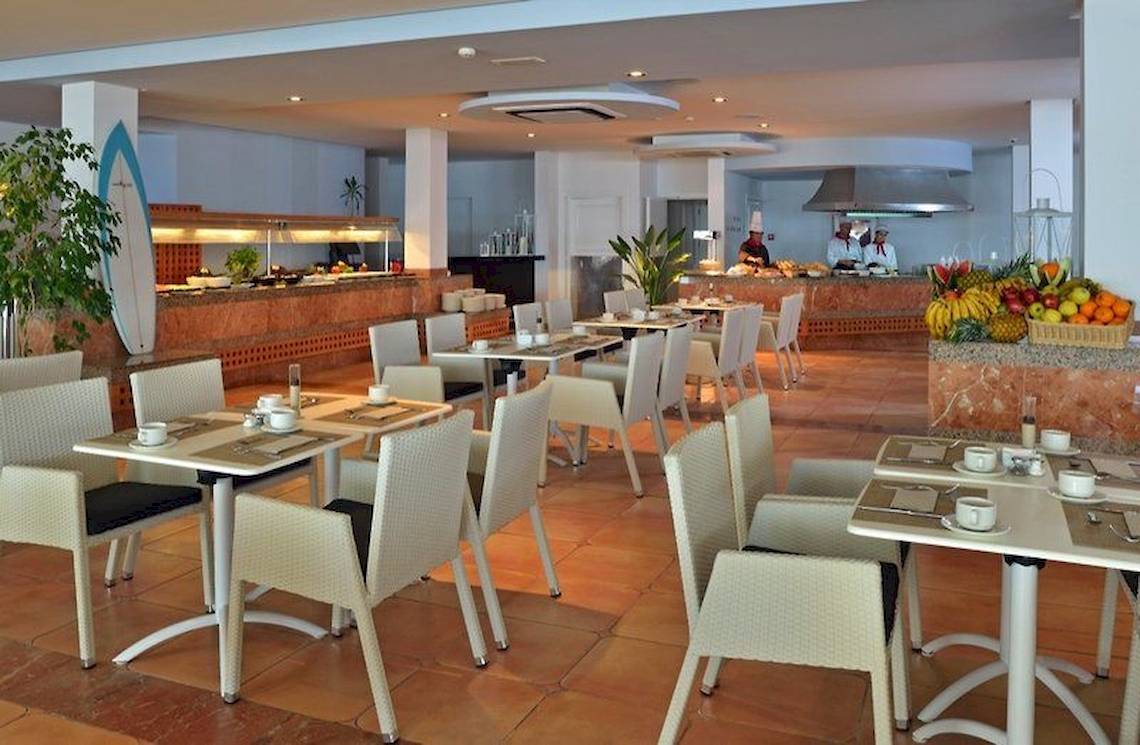 Restaurant des Sol Wave House All Suites mit vielen Tischen und Stühlen, serviertem Frühstück und Köchen im Hintergrund.