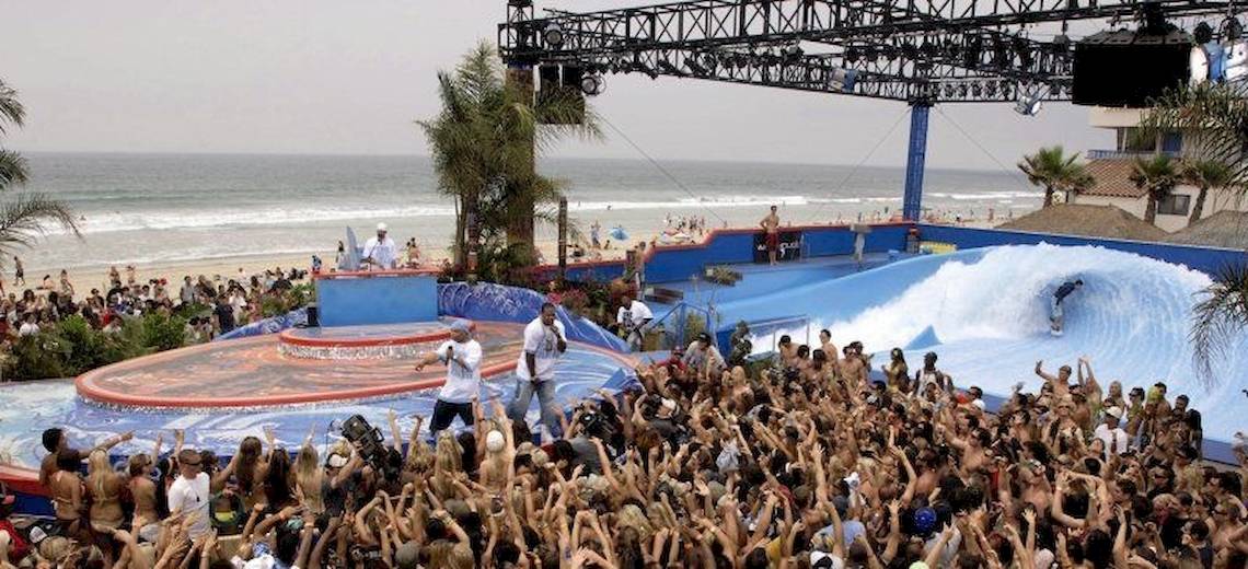 Eine Menschenmenge auf dem Strand beobachtet ein Surfereignis, ein Surfer reitet auf einer künstlichen Welle.