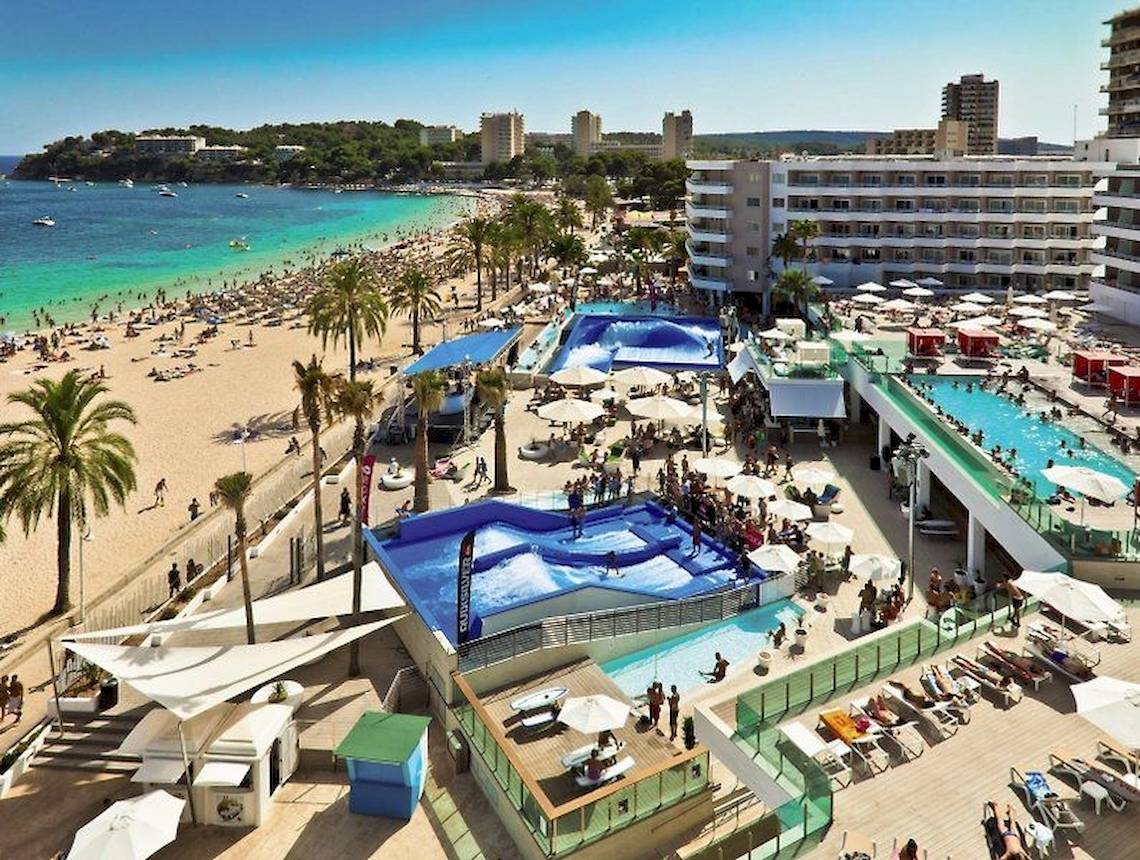 Luftaufnahme eines Strandes mit Hotel und Wellenpool, viele Besucher, Palmen und Sonnenschirme.