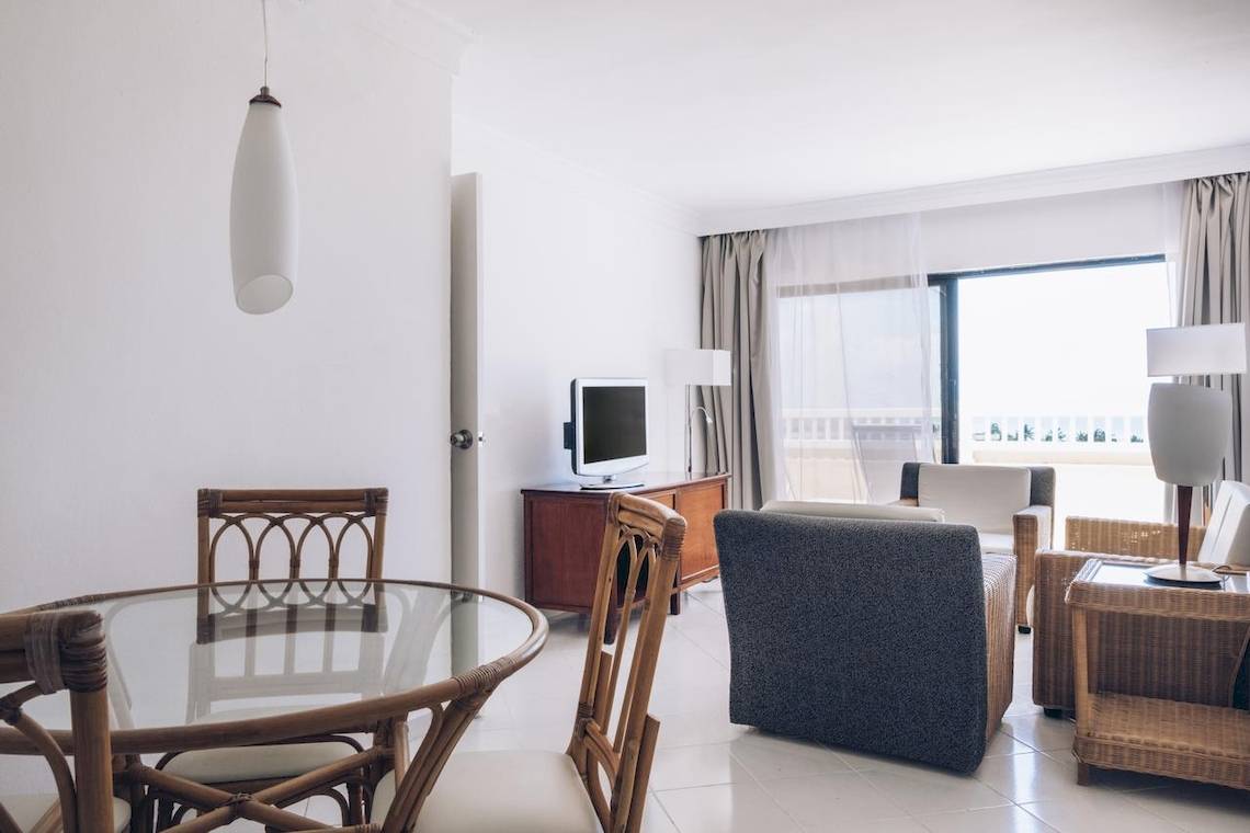 Innenansicht eines Hotelzimmers im Iberostar Origin Bella Costa mit Tisch, Stühlen, Fernseher und großem Fenster.