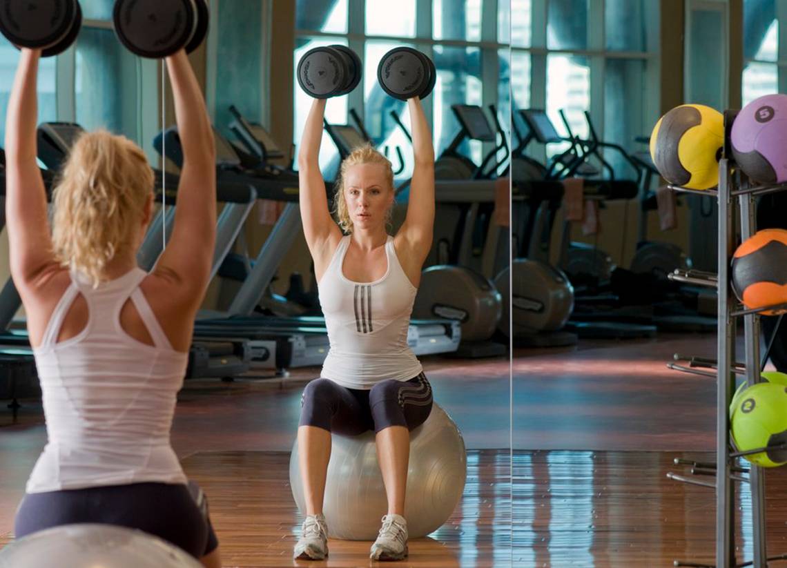 Zwei Frauen im Fitnessstudio machen Übungen mit Hanteln auf einem Gymnastikball.
