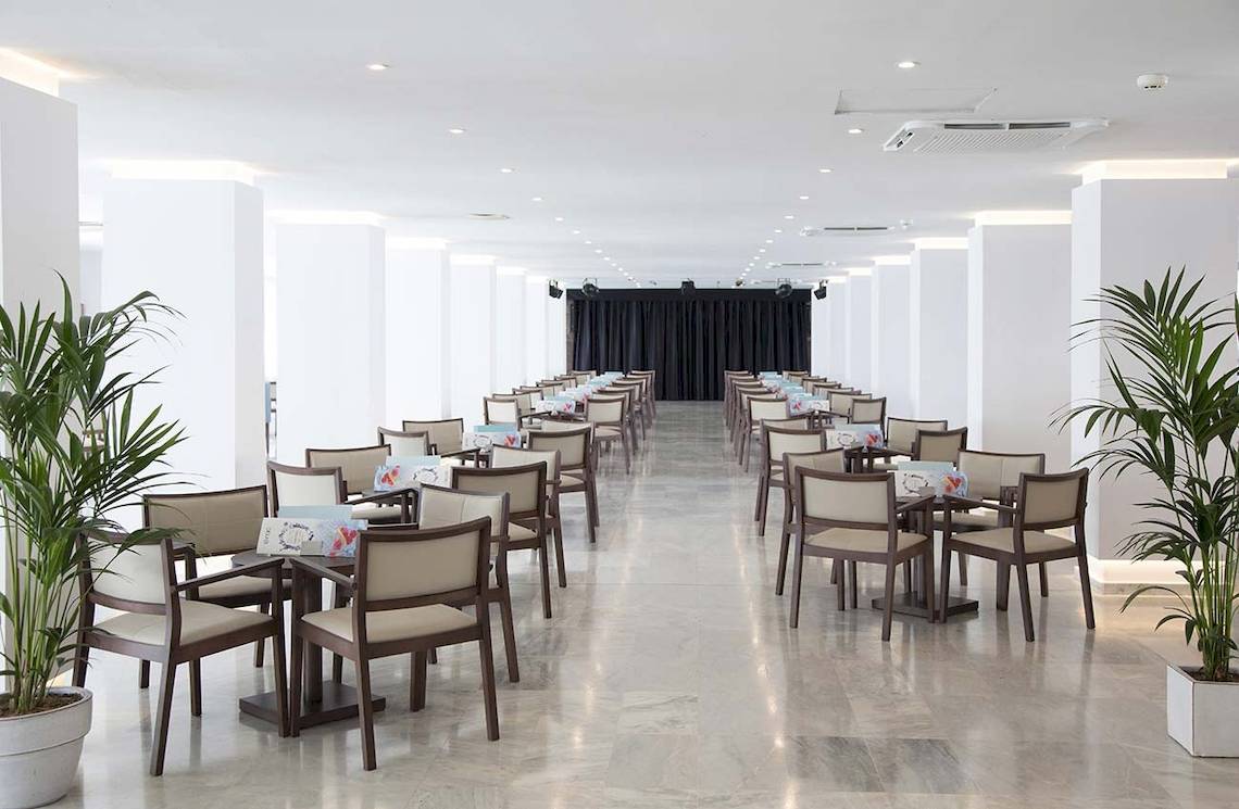 Innenansicht des Speisesaals im Alua Leo Hotel mit geräumigen weißen Wänden, braunen Stühlen und Tischen, sowie grünen Pflanzen in Töpfen.