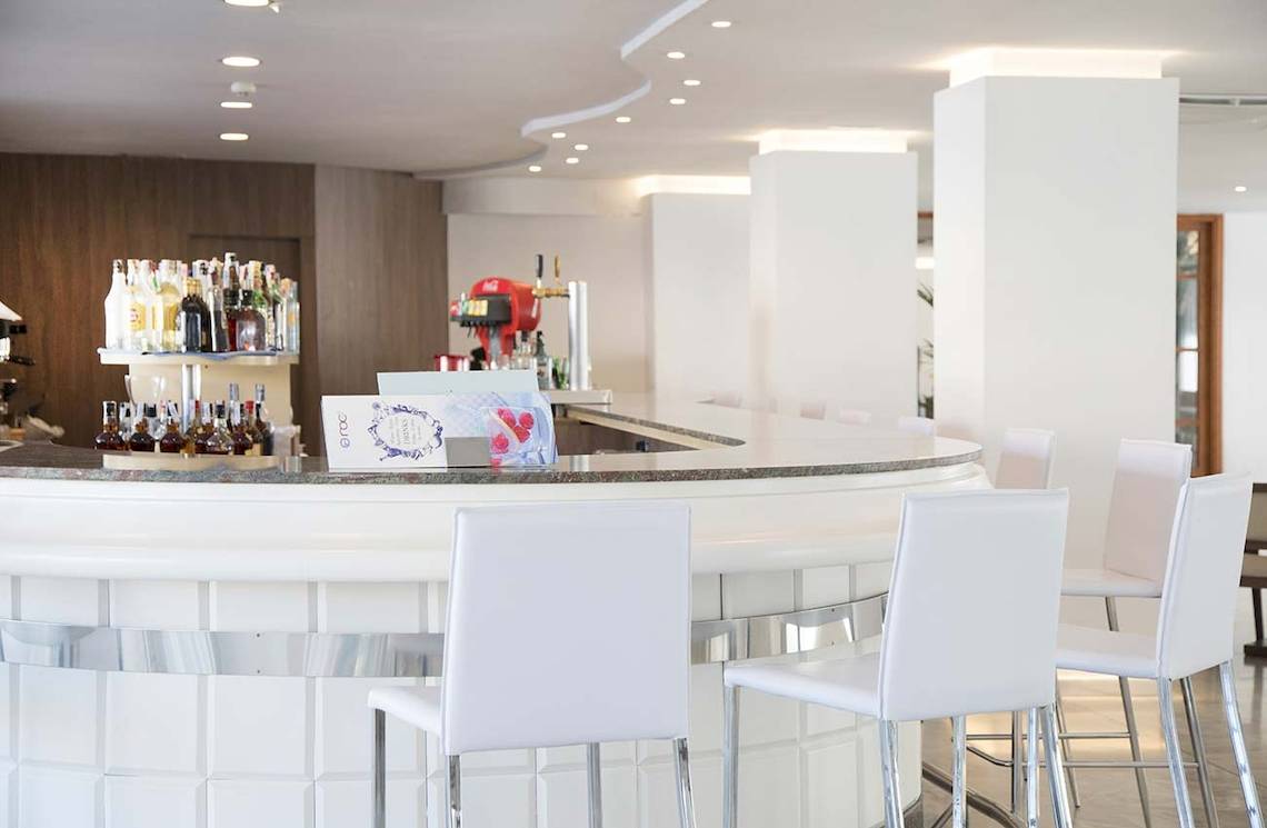 Barbereich im Alua Leo Hotel mit weißen Stühlen und einer Theke voller Flaschen.