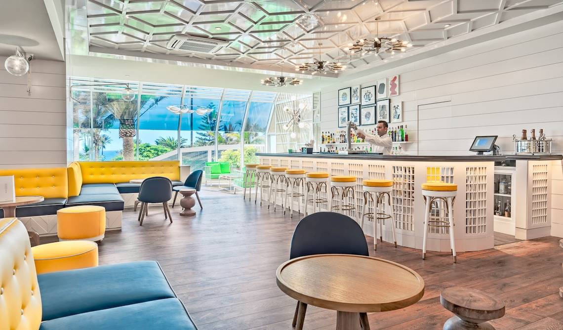 Innenansicht der Bar im H10 Las Palmeras Hotel mit Barkeeper, gelben Stühlen und modernem Design.