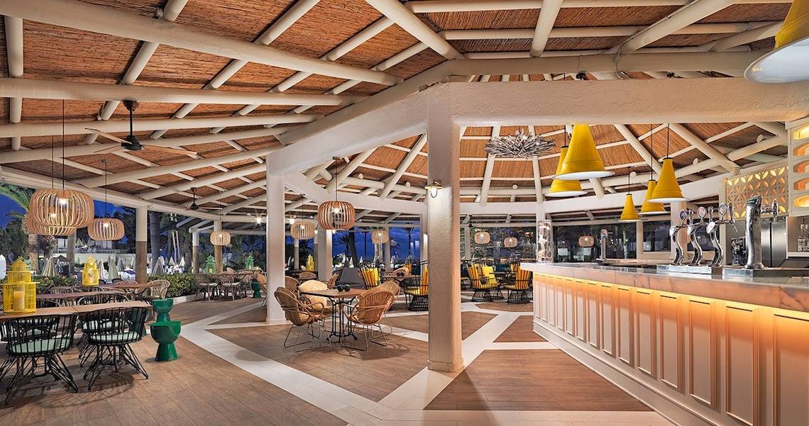 Innenbereich des H10 Las Palmeras Hotels mit Bar, Tischen und Stühlen, gelben Lampen und Holzboden.
