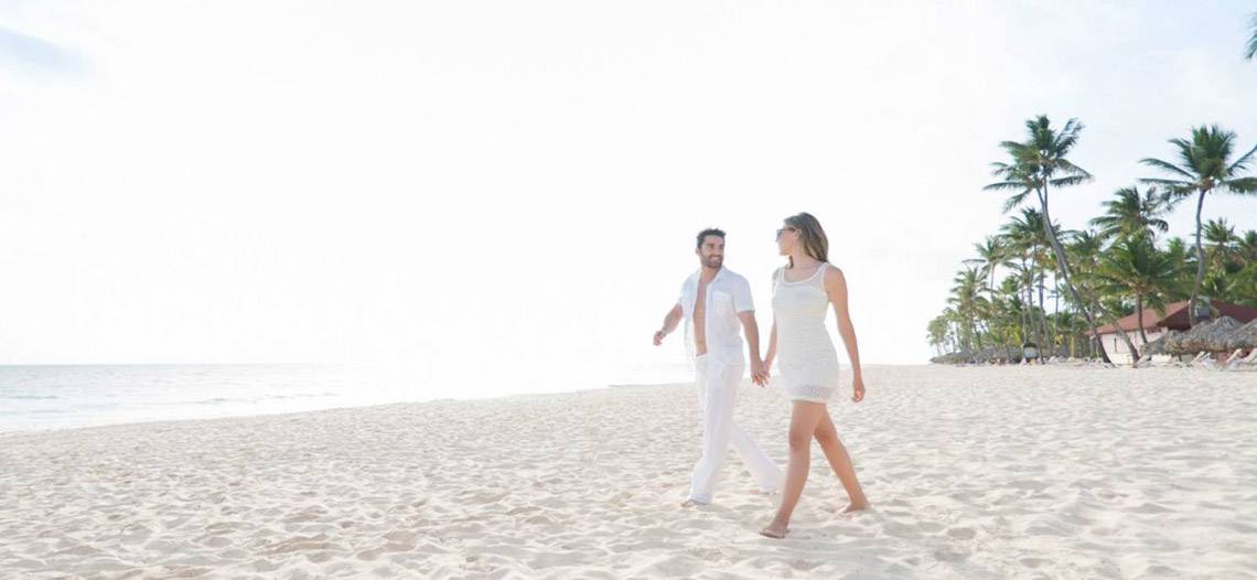 Ein Paar, das Hand in Hand auf einem weißen Sandstrand läuft, mit Palmen und Gebäuden im Hintergrund.